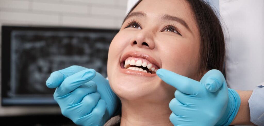 A dentist wearing blue gloves examines a smiling patient's teeth during a dental consultation. The patient reclines in a dental chair while the dentist points to her smile, highlighting cosmetic dentistry, oral health assessment, and personalized dental care.