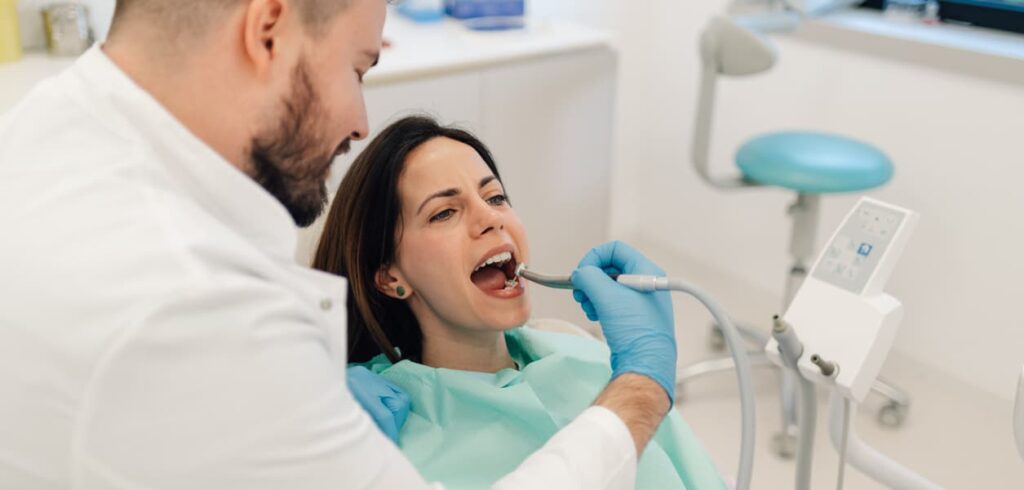 A dentist wearing gloves performs a routine dental examination on a patient seated in a modern dental chair. The bright treatment room features contemporary dental equipment, highlighting preventive oral care and professional dental services.