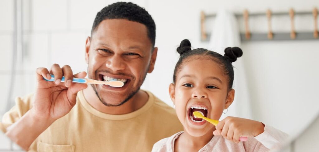 A father and his young daughter smile while brushing their teeth together in a bright bathroom. The image highlights healthy oral hygiene habits, family dental care, and teaching children the importance of brushing their teeth daily.
