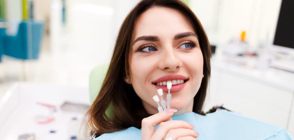 A patient in a dental chair compares tooth shade samples to her smile before a cosmetic dental treatment. The image represents professional teeth whitening, smile makeovers, and personalized cosmetic dentistry to achieve natural-looking results.