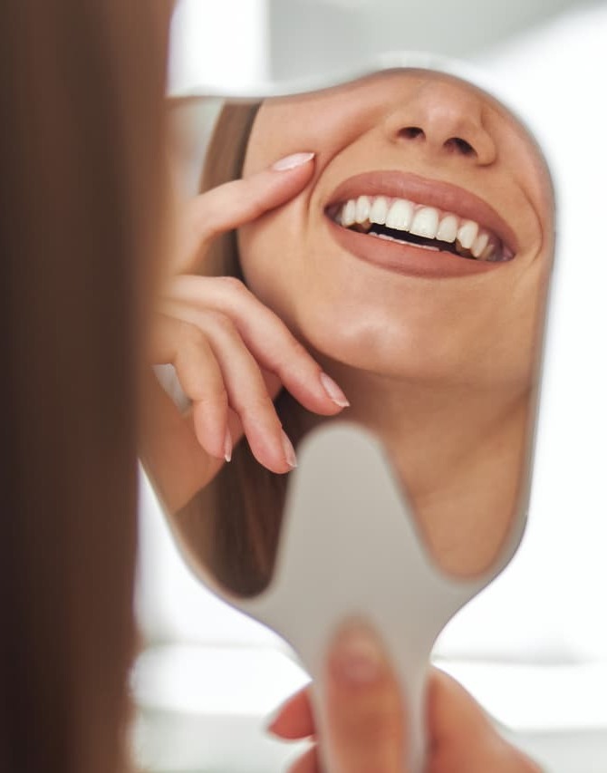 A patient smiles while looking into a handheld mirror after a cosmetic facial treatment, admiring refreshed, natural-looking results. The image represents confidence, facial rejuvenation, and satisfaction with non-surgical aesthetic care.