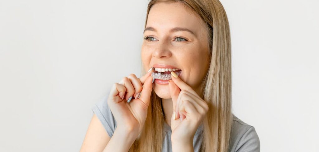 A smiling woman inserts a clear dental aligner onto her upper teeth using both hands. The close-up image highlights clear aligner orthodontic treatment, straightening teeth, and at-home dental care against a clean, neutral background.